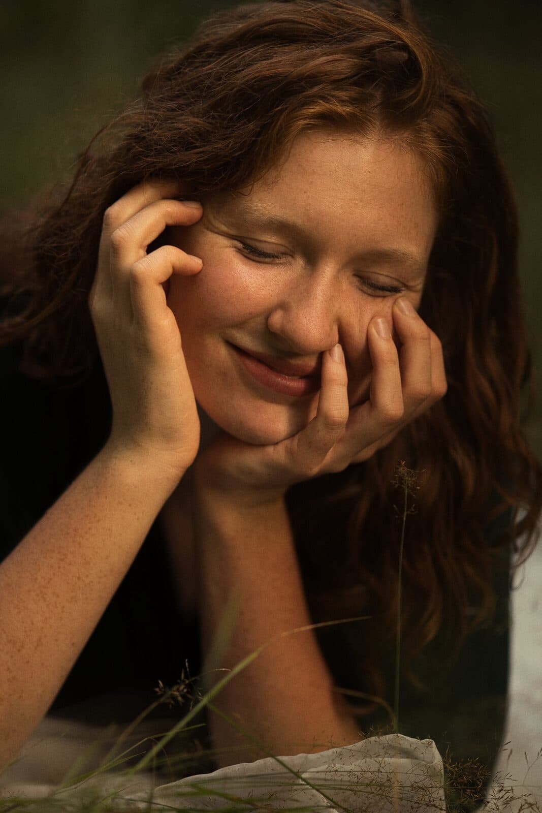 Red-haired woman smiling with her face resting on her hands in the grass